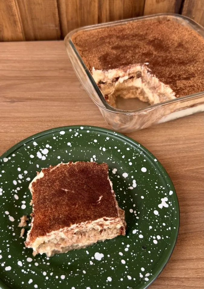 Imagen de un tiramisú en un plato verde con una fuente con el tiramisú entero en el fondo.